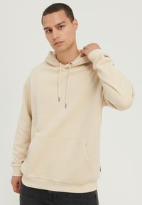 Sudadera con capucha beige hecha de tela suave con un bolsillo frontal, cordones y puños acanalados. Diseño sencillo y de ajuste relajado.