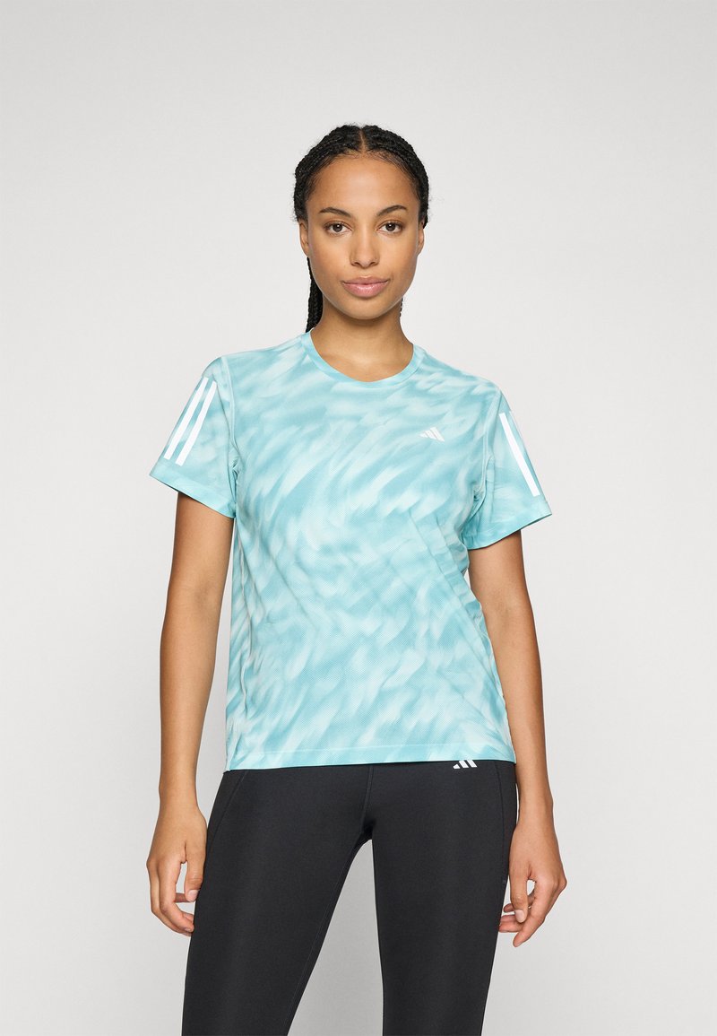 Donna con capelli intrecciati, che indossa una maglietta sportiva leggera blu chiaro a fantasia e leggings neri, in piedi contro uno sfondo bianco uniforme.