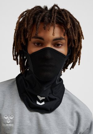 Person mit mittellangen Dreadlocks, der eine schwarze Netz-Gesichtsmaske und einen grauen Hummel-Pullover vor einem einfachen Hintergrund trägt.