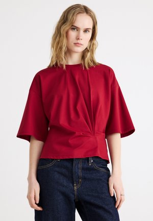 Femme aux cheveux blonds ondulés portant un haut rouge ample à manches courtes avec un twist à l'avant et un jean bleu foncé, debout devant un fond blanc.