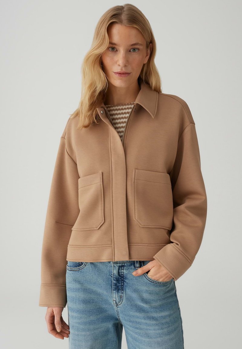 Veste courte beige avec col à revers, poches avant et texture lisse, associée à un jean en denim bleu clair.