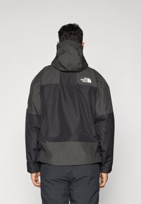 Muž oblečený v čierne a šedé bundy The North Face s kapucňou, zobrazený zozadu na jednoduchom svetlom pozadí.