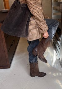 Osoba w brązowych zamszowych butach i kraciastej kurtce zakładająca jeden but, wewnątrz, obok kuchennych szafek i metalowych półek.