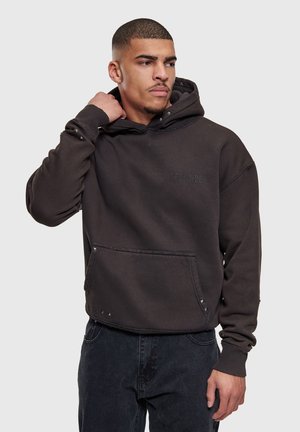 Jonge man met kort haar, gekleed in een donker oversized hoodie met studs, houdt de capuchon bij zijn nek vast en kijkt opzij.