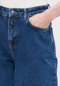 Modré džíny s vysokým pasem, předními kapsami a kontrastním žlutým šitím. Tkanina je strukturovaná s klasickým designem džínů.