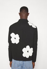 Chaqueta negra con diseños florales blancos en la parte trasera y las mangas. Presenta un cuello y mangas largas, hecha de un tejido texturizado.