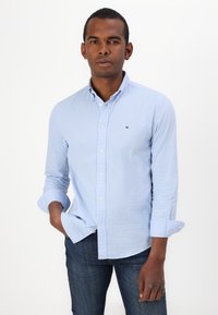 Camicia a quadri blu chiaro con maniche lunghe, bottoni bianchi e un piccolo logo sul petto, abbinata a un paio di jeans di denim scuro.