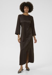 Kvinna med lockigt hår som bär en lång mörkbrun klänning med 3/4 ärmar, slits fram, beige armband och zebramönstrade klackar mot en enfärgad bakgrund.