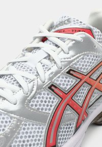 Λευκό αθλητικό παπούτσι με ασημί πλέγμα επάνω, κόκκινες και μαύρες λεπτομέρειες, και λευκές κορδόνια. Διαθέτει υφή στην επιφάνεια και μεσαίο πάτο με επένδυση.