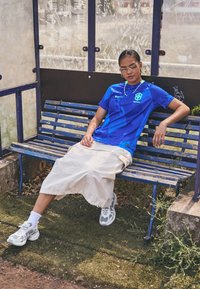 Blue Nike jersey with graphic design, beige knee-length skirt, white socks, and gray sneakers, seated on a blue bench in an outdoor setting.