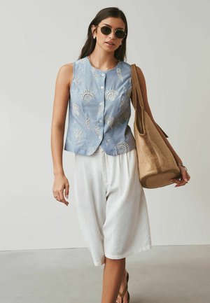 Woman wearing round sunglasses, sleeveless blue top with white pattern, white knee-length skirt, carrying large tan woven tote bag on shoulder.