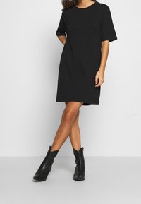 Nainen pukeutuneena yksinkertaiseen mustaan lyhythihaiseen polvipituiseen mekkoon ja mustiin nilkkasaappaisiin, seisoo ristitettyjä jalkoja vasten valkoista taustaa.