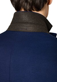 Granatowa kurtka z teksturowanej tkaniny, z ciemno-brązowym filcowym kołnierzem. Zbliżenie na plecy, pokazujące szwy i szczegóły kołnierza.