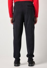 Osoba oblečená v černých cargo teplácích a černých teniskách, stojící zády k fotoaparátu, s dlouhým červeným tričkem.