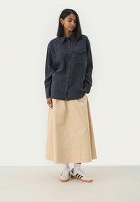 Marineblauwe button-up shirt met een zak, gecombineerd met een beige enkellange rok. Model draagt witte sneakers met zwarte accenten en enkelsokken.