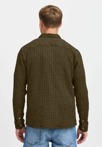 Camisa verde texturizada con un patrón de gofre, de manga larga, con botones y cuello, llevada sobre unos jeans azul claro.