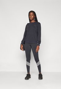 Mørkegrå hettegenser med ribbestrikkede mansjetter og kant, kombinert med tettsittende svarte leggings med grå chevron-detaljer på leggene. Svarte joggesko.