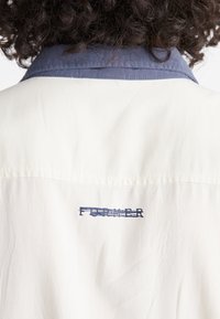 Camicia di un leggero color crema con colletto blu navy, caratterizzata da un ricamo testurizzato "FOREVER" sulla parte posteriore. Tessuto morbido con una leggera lucentezza.