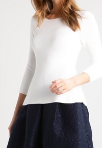 Weißes, geripptes Stricktop mit dreiviertel Ärmeln, verziert mit einem eng anliegenden Oberteil und einem ausgestellten Saum. Kombiniert mit einem dunklen, strukturierten Rock.