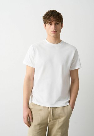 Witte katoen t-shirt met korte mouwen, ronde hals en een gladde textuur. Model draagt het in combinatie met beige cargo broek en staat met één hand in de zak.