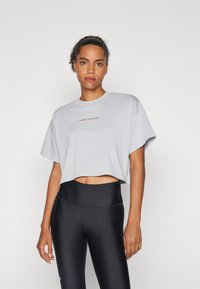 Sivi skraćeni majica s roliranim kratkim rukavima, s "Under Armour" logotipom u crvenoj boji na teksturiranoj površini, u kombinaciji s crnim tajicama visokog struka.