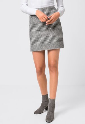 Kobieta ubrana w szarą plisowaną mini spódniczkę w kratę, biały prążkowany top z długim rękawem oraz szare zamszowe botki, stoi na jednolitym tle.