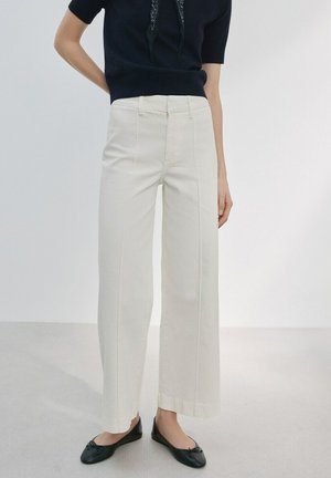 Donna che indossa pantaloni avorio a vita alta e gamba larga, top nero a maniche corte, ballerine nere, in piedi contro uno sfondo chiaro e uniforme.