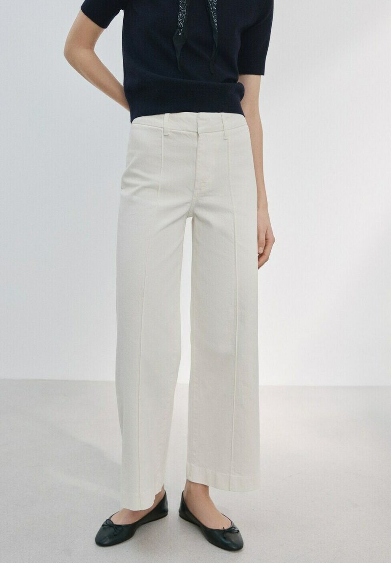 Frau trägt hoch taillierte, weit geschnittene, cremeweiße Hose, schwarzes Kurzarmoberteil, schwarze Ballerinas, steht vor einem einfarbigen hellen Hintergrund.