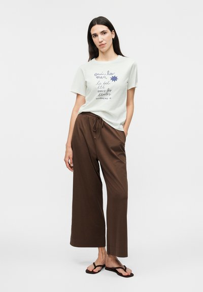 Γυναίκα με ίσια σκούρα μαλλιά, που φοράει λευκό μπλουζάκι με σχέδιο, καφέ φαρδιά παντελόνια και μαύρα σαγιονάρες, στέκεται με το ένα χέρι στην τσέπη.