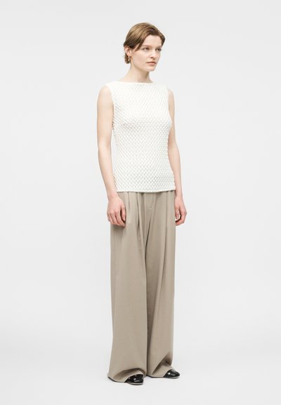 Γυναίκα με λευκό υφασμάτινο αμάνικο τοπ, φαρδιά μπεζ παντελόνια, μαύρα παπούτσια και μικρά χρυσά κρίκους σκουλαρίκια, στέκεται μπροστά σε λευκό φόντο.