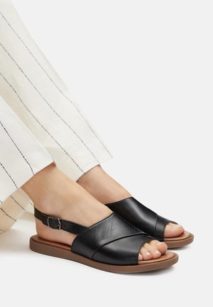 Sandalias negras de cuero con diseño de correas cruzadas, punta abierta y correa en el tobillo. Acabado texturizado, combinadas con pantalones de rayas claras.