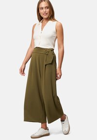 Olive green wide-leg pants with a tied waist detail, paired with a cream ribbed sleeveless top. Model stands in white sneakers.