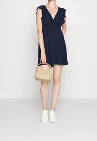 Gaun eyelet biru tua dengan lengan pendek berkerut, desain v-neck, pinggang yang pas, dan rok melebar, dipadukan dengan tas anyaman beige dan sepatu sneakers putih.