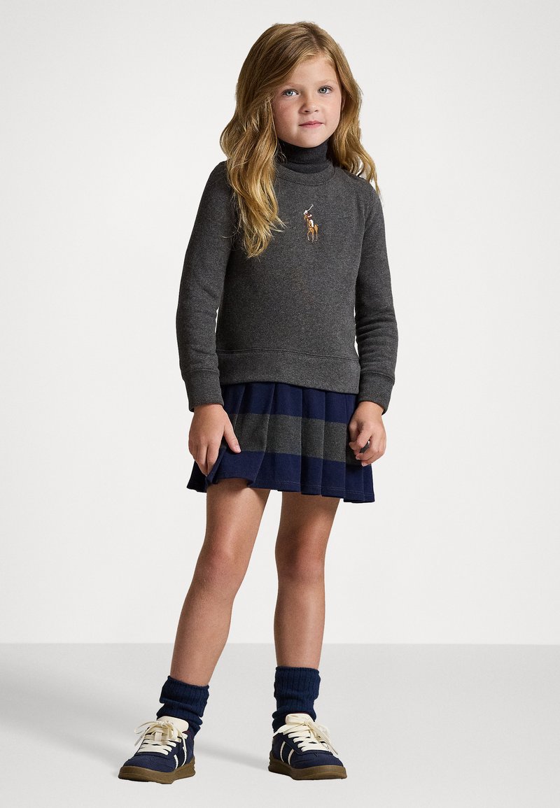 Suéter cinza de gola alta com logotipo bordado, emparelhado com uma saia plissada listrada em azul escuro e cinza. Ténis marinhos com atacadores em creme.