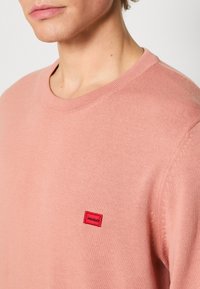 Gros plan sur une personne portant un pull ras du cou rose clair avec un petit logo rectangulaire rouge "HUGO" sur la poitrine.