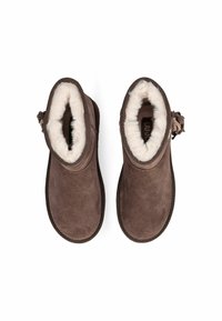 Stivali ankle in suede marrone con fodera in pelliccia bianca, punta arrotondata e una cinghia decorativa laterale con accenti in metallo.