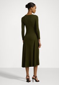 Olivgrön långärmad klänning med åtsittande kropp och utsvängd kjol, tillverkad av ett smidigt tyg. Anmärkningsvärd detalj på ryggen och längd ovanför anklarna.