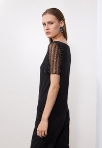 Top scurt negru cu mâneci transparente și brodate. Țesătură netedă, decolteu rotund și siluetă ajustată, asortat cu pantaloni negri.