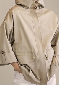 Beżowa wodoodporna kurtka z kapturem, dużymi kieszeniami i guzikowymi zdobieniami. Charakteryzuje się luźnym krojem i podwiniętymi rękawami. Gładka faktura.