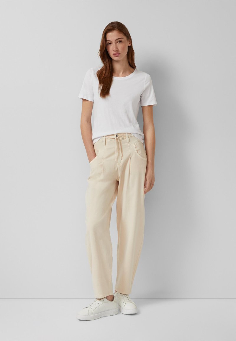 Jeune femme portant un t-shirt blanc à manches courtes, un pantalon beige taille haute avec cordon de serrage, des baskets blanches, debout les mains dans les poches.