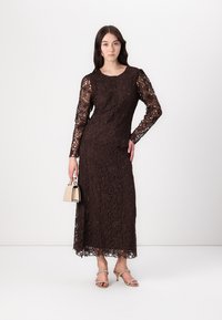 Braunes Spitzenkleid mit langen Ärmeln, floralen Mustern, gerader Silhouette und gewelltem Saum, kombiniert mit einer kleinen Handtasche und Riemchenschuhen.