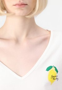 T-shirt blanc à col en V avec une illustration de citron jaune et des feuilles vertes, avec le texte "Squeeze the day" sur le côté gauche.