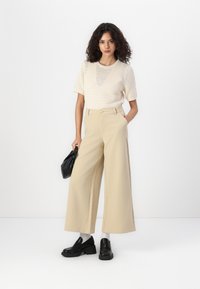 Gebreid crèmekleurig topje met korte mouwen, gecombineerd met beige wijde broek. Zwarte chunky schoenen en een zwarte handtas maken de look af.
