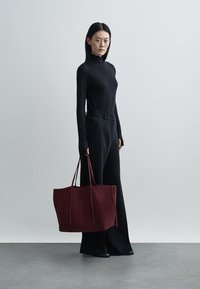 Large burgundy suede tote bag with long handles, paired with a black long-sleeve top and wide-leg black trousers. Smooth texture.