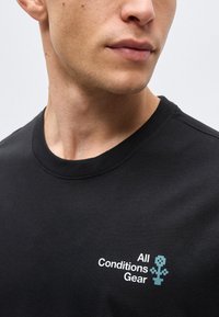 Άνδρας που φοράει μαύρο μπλουζάκι με λαιμόκοψη και το κείμενο «All Conditions Gear» μαζί με ένα ψηφιοποιημένο μπλε λουλούδι στο αριστερό στήθος.