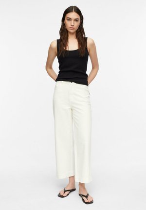 Woman stands with hands behind back wearing black sleeveless top, white wide-leg pants, and black flip-flop sandals on white background.