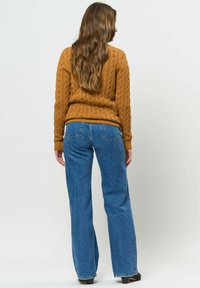 Femme aux longs cheveux bruns portant un pull moutarde en maille torsadée, un jean bleu droit et des chaussures à talons noires, debout dos à nous.