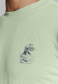 Lichtgroene t-shirt met korte mouwen van een zachte stof, met een afbeelding van een klassieke buste met een duikbril en de tekst "HET IS ISALY, ROMA."