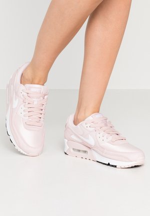 Beine, die hellrosa Nike Air Max Sneaker mit weißen Swoosh-Logos tragen, stehend auf einem schlichten hellgrauen Boden und Hintergrund.