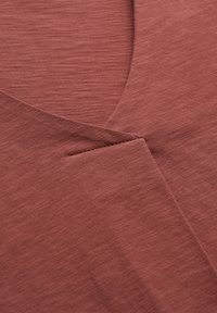 Rustfarvet stof med en tekstureret overflade og et V-hals design. Indeholder overlappende forsektioner og diskrete syninger.